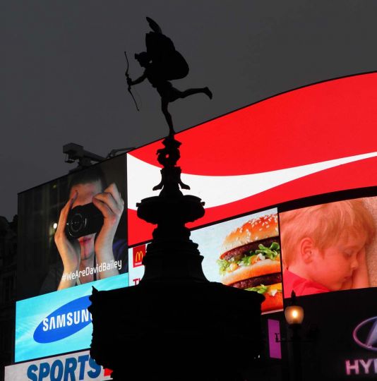 Anteros figure silhouetted against floodlit adverts panels, Piccadilly Circus, London, UK. The statue of the nude winged archer, at the top of The Shaftesbury Monument Memorial Fountain, is known as Eros Column, 1892-1893, aluminium cast, by Alfred Gilbert. © Manuel Cohen