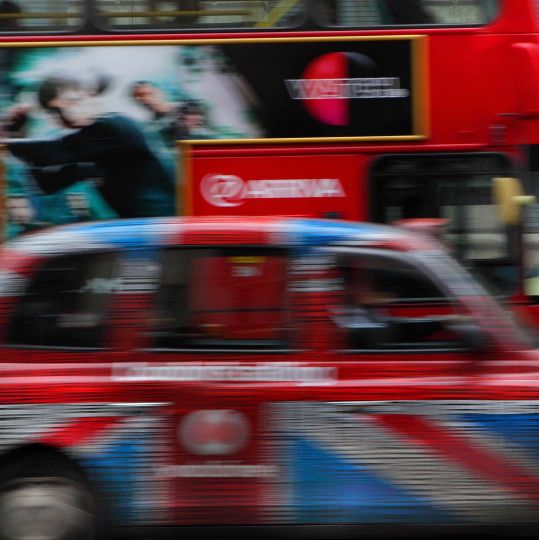 High Speed London cab, colored with royal banner Union Jack (United Kingdom flag), against a red London bus with adverts, London, UK. © Manuel Cohen