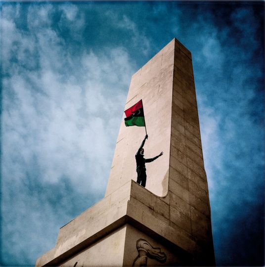 © Michael Christopher Brown, LIBYA. Benghazi. March 2, 2011. 15:30:56. A young man waves the  ag of the newly created Libyan Repub- lic (and the former  ag of the Kingdom of Libya) atop Nasser statue during the burning of Gadda ’s Green Books, near the Green Book Museum.