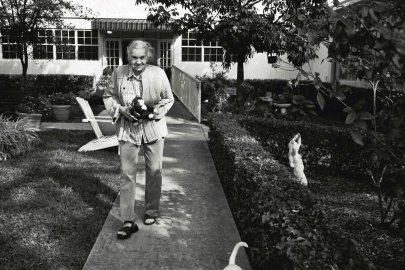 Madje Steber walks to the dining room for lunch at Bay Oaks, a small beautiful assisted living facility, along with one of the facility's pet dogs, a week after being moved from her home in Austin, Texas to Miami, Florida, to be closer to her daughter, Maggie. She carries her favorite stuffed toy. A scientist all her life, Madje began to lose her memory at the age of 78. An eccentric recluse, Madje blossomed and became very socialable once she moved into this small facility where the caregivers took special care of everyone. © Maggie Steber - Courtesy Leica Gallery Los Angeles