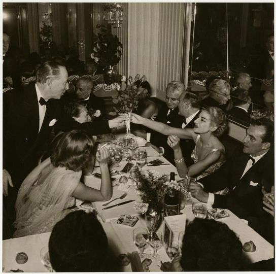Maria Callas receives a bouquet of flowers from Valentino Bompiani at the Savini restaurant on the evening of the premiere of Gaspare Spontini's La Vestale. To her left is director Luchino Visconti, at the head of the table is her husband Giovanni Battista Meneghini, to the left of whom sits the superintendent of Teatro alla Scala, Antonio Ghiringhelli. Milan, 1954. Photograph by Franco Giglio – Publifoto, 23 x 23.20 cm; Publifoto Archive (Callas_002); Maria Callas. Portraits from the Intesa Sanpaolo Publifoto Archive; Gallerie d’Italia – Milano, Intesa Sanpaolo Museum