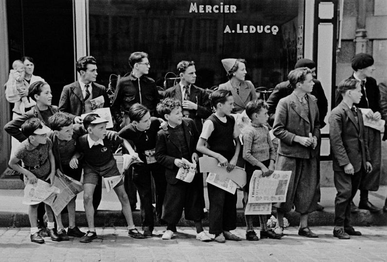 A crowd gathered in front of Mr. Pierre Cloarec's bicycle shop. The owner of the shop is racing in the Tour de France, Pleyben, Brittany, France, July 1939. © Robert Capa © International Center of Photography/Magnum Photos
