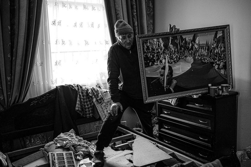 Ivan stands in a bedroom of his Andriivka summer house, invaded and ransacked by Russian troops during their four-week village occupation. He shows a picture prominently featuring his face sliced by the soldiers, made ten years ago in Kyiv during a Ukrainian protest against the Russian language law. Living in Kyiv, he thought about escaping to this house for safety during the start of the invasion. April, 2022. © Byron Smith - From the book Testament ’22 - Courtesy Verlag Kettler